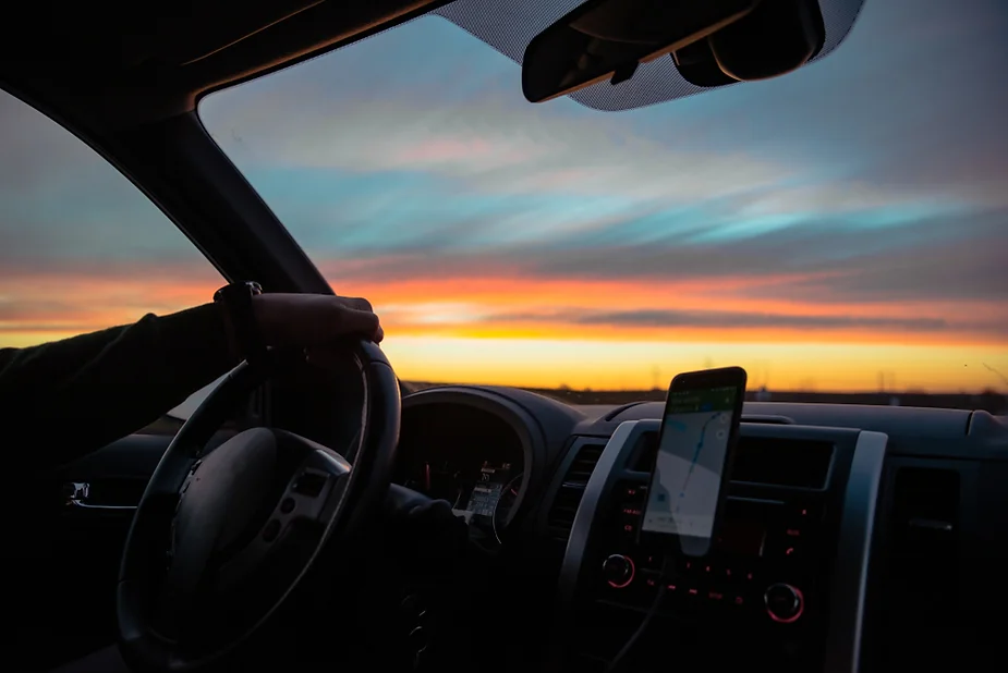 Gasolineras en Islandia Una mano sujeta un volante de un vehículo en marcha mientras a través de la luna del coche se observa un atardecer.