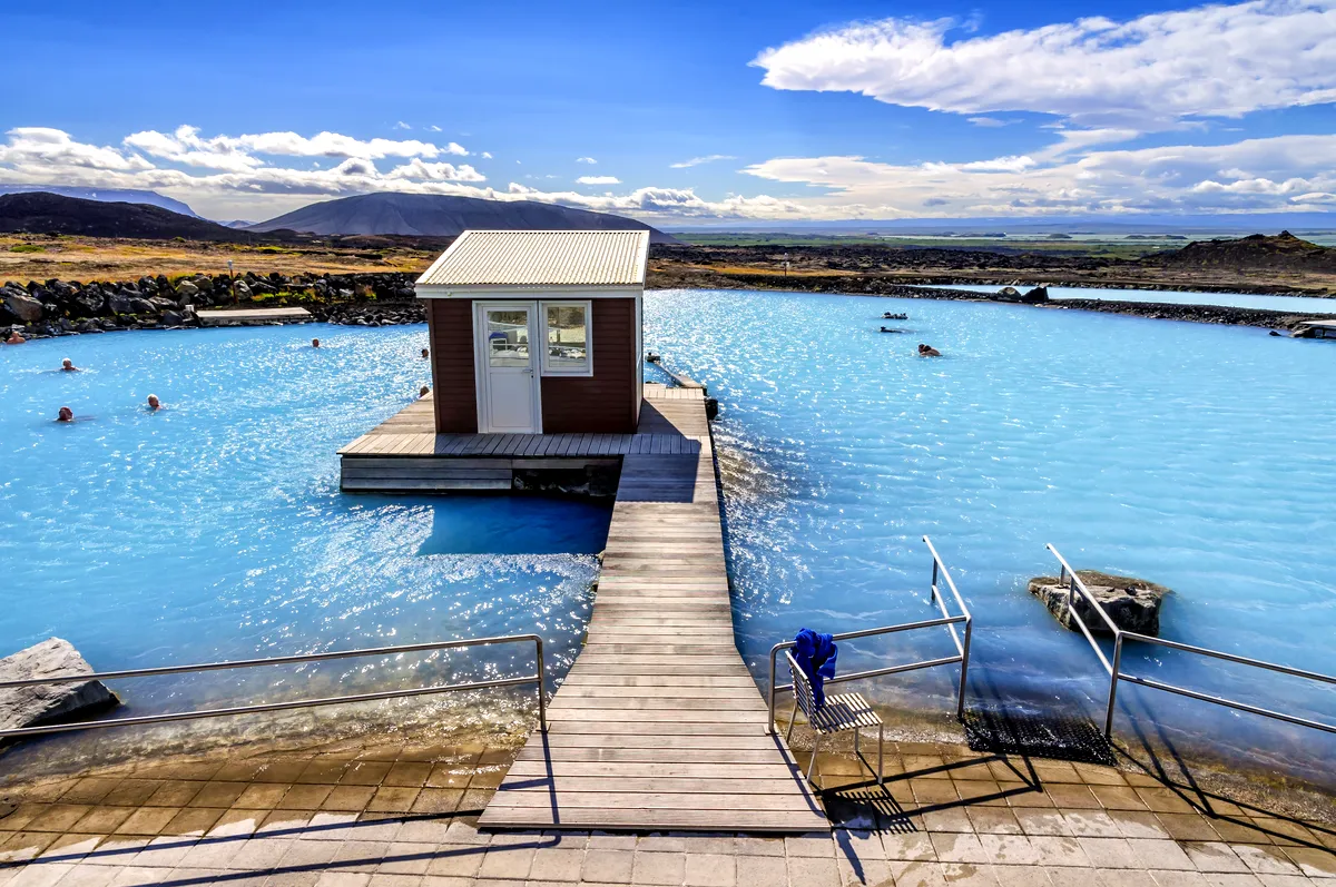 Laguna geot&eacute;rmica de Myvatn bajo un cielo despejado, con aguas azules brillantes que reflejan la luz del sol. La imagen captura el peque&ntilde;o edificio central rodeado de piscinas, con monta&ntilde;as y un vasto paisaje esc&eacute;nico de fondo