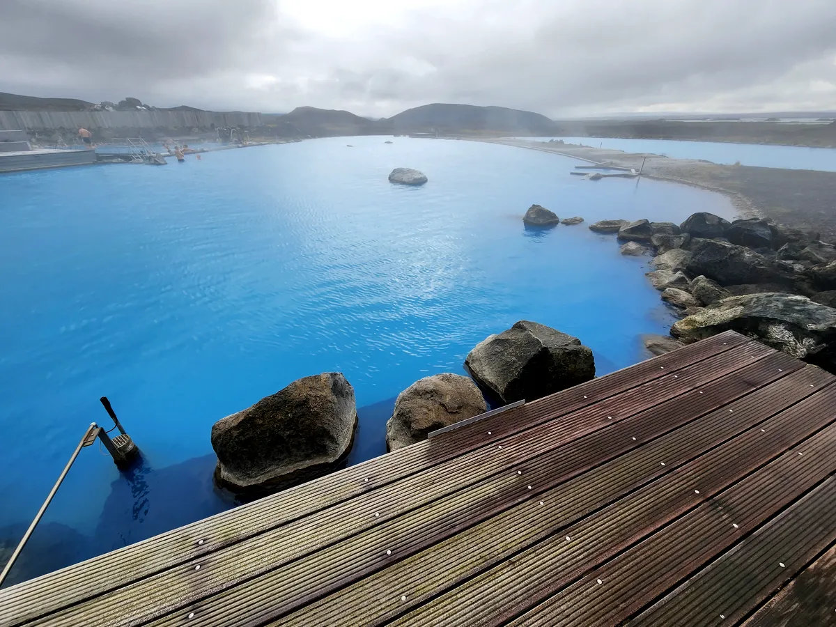 Una vista m&aacute;s cercana de las tranquilas aguas azules de la laguna geot&eacute;rmica de Myvatn en Islandia. Las rocas y las pasarelas de madera crean una atm&oacute;sfera tranquila mientras el vapor se eleva desde las c&aacute;lidas aguas, mientras que el paisaje lejano, envuelto en niebla, a&ntilde;ade un toque misterioso al entorno.