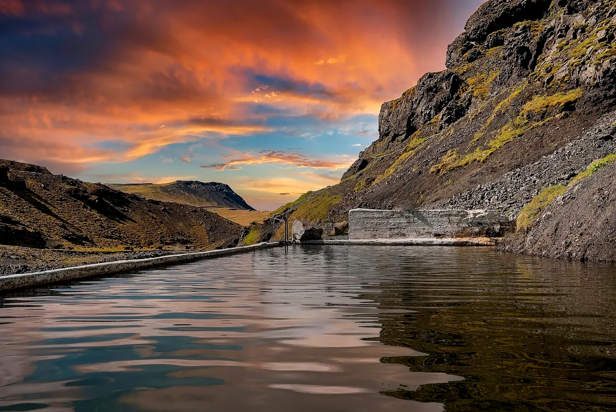 Una puesta de sol pac&iacute;fica ba&ntilde;a las escarpadas monta&ntilde;as y las tranquilas aguas de las aguas termales de Seljavallalaug con colores c&aacute;lidos. El reflejo en la superficie tranquila se suma a la belleza surrealista del paisaje island&eacute;s