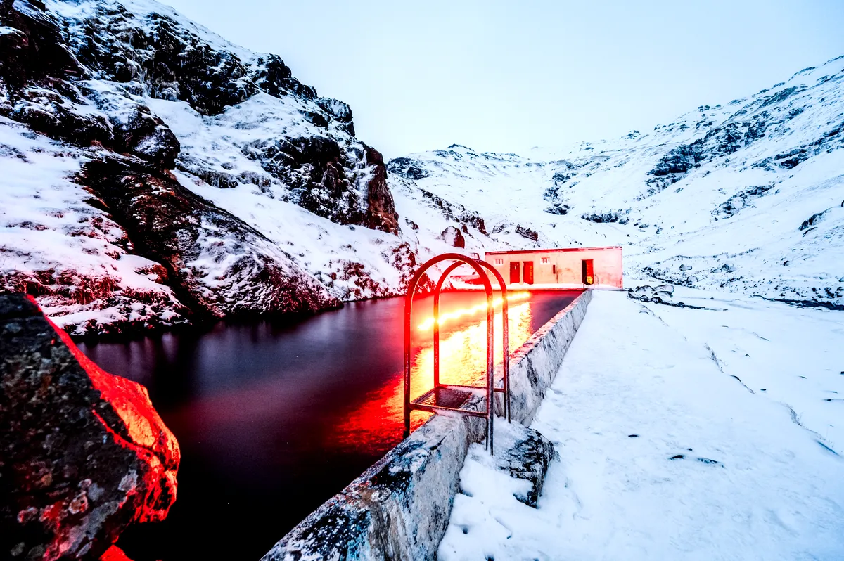 Una serena piscina geotermal enclavada entre las laderas nevadas de las monta&ntilde;as de Seljavallalaug, Islandia. La tenue iluminaci&oacute;n le da a la escena un brillo m&iacute;stico, que se refleja en las rocas cubiertas de nieve.