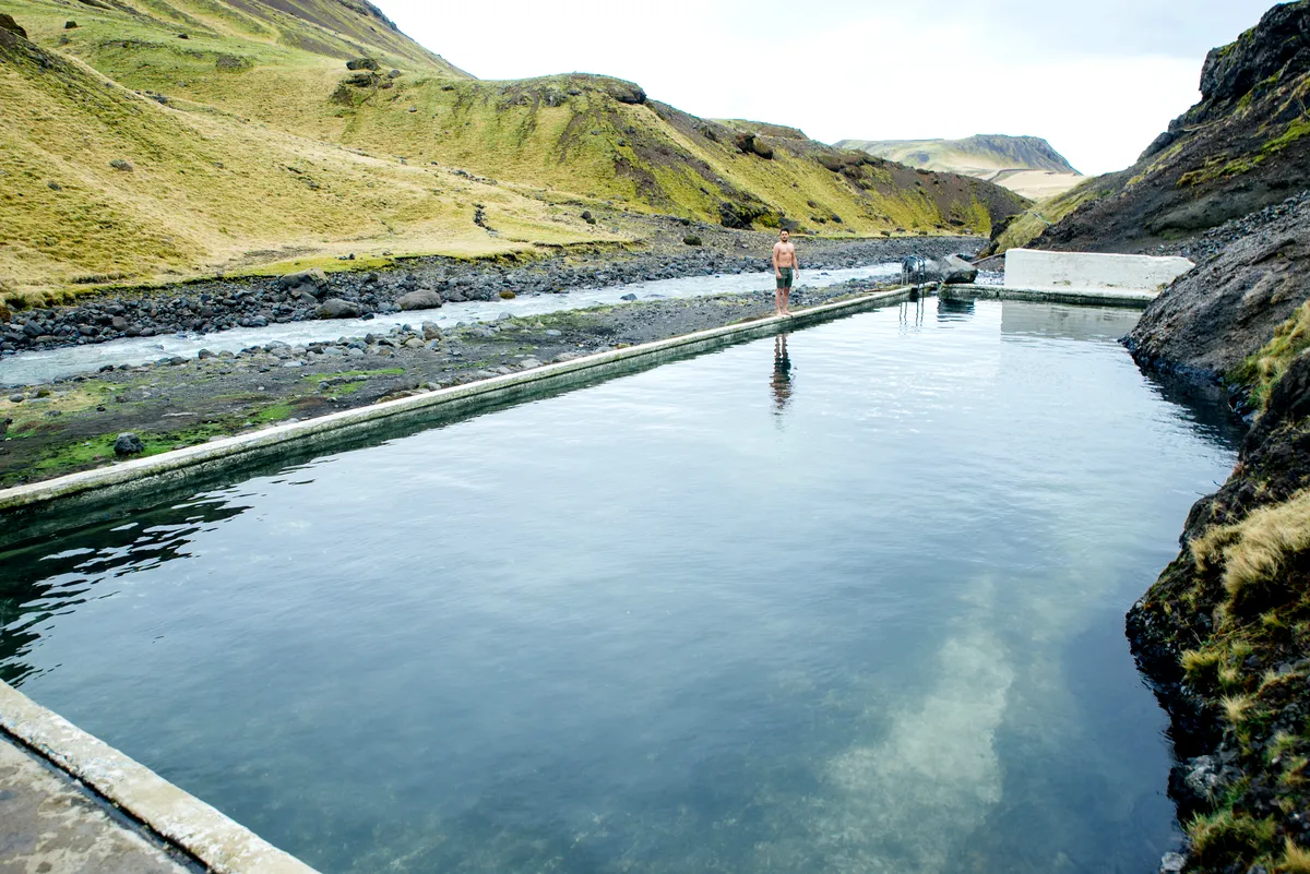 Un hombre se encuentra cerca del borde de las aguas cristalinas de Seljavallalaug piscina geot&eacute;rmica, rodeada de colinas cubiertas de musgo. El paisaje natural ofrece un escape sereno y remoto en Islandia.