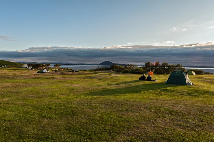 Un camping esc&eacute;nico en Islandia con tiendas de campa&ntilde;a sobre c&eacute;sped verde, con vistas a un lago tranquilo y monta&ntilde;as a lo lejos, ilustrando la belleza de los campings designados en Islandia.