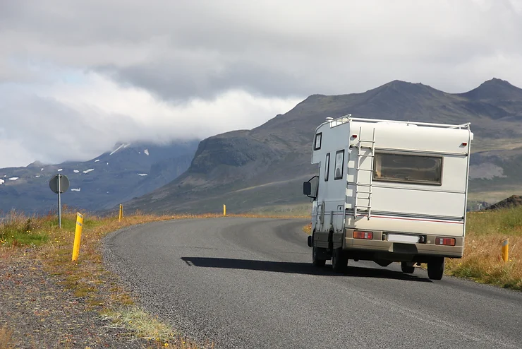 Una campervan circulando por una carretera esc&eacute;nica en Islandia, rodeada de monta&ntilde;as y paisajes dram&aacute;ticos, destacando la libertad y aventura de viajar en campervan por la isla.