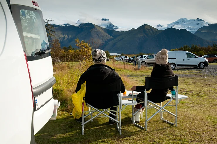 Dos viajeros disfrutando de un camping esc&eacute;nico en Islandia, sentados fuera de su campervan con vistas a monta&ntilde;as y glaciares. Un ejemplo perfecto de un camping bien equipado para viajeros en campervan.