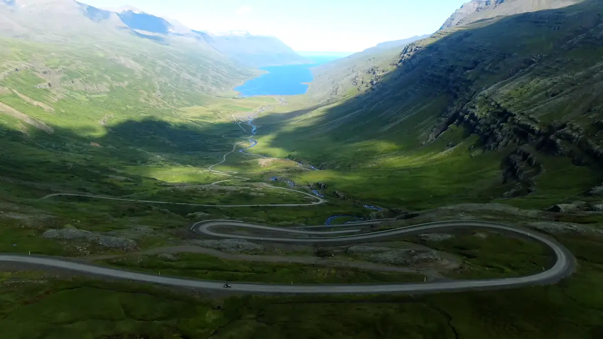 Carretera serpenteante a través de un valle verde en los Fiordos del Este de Islandia, con un fiordo azul al fondo. Conducir una autocaravana permite explorar estos paisajes remotos a tu propio ritmo.