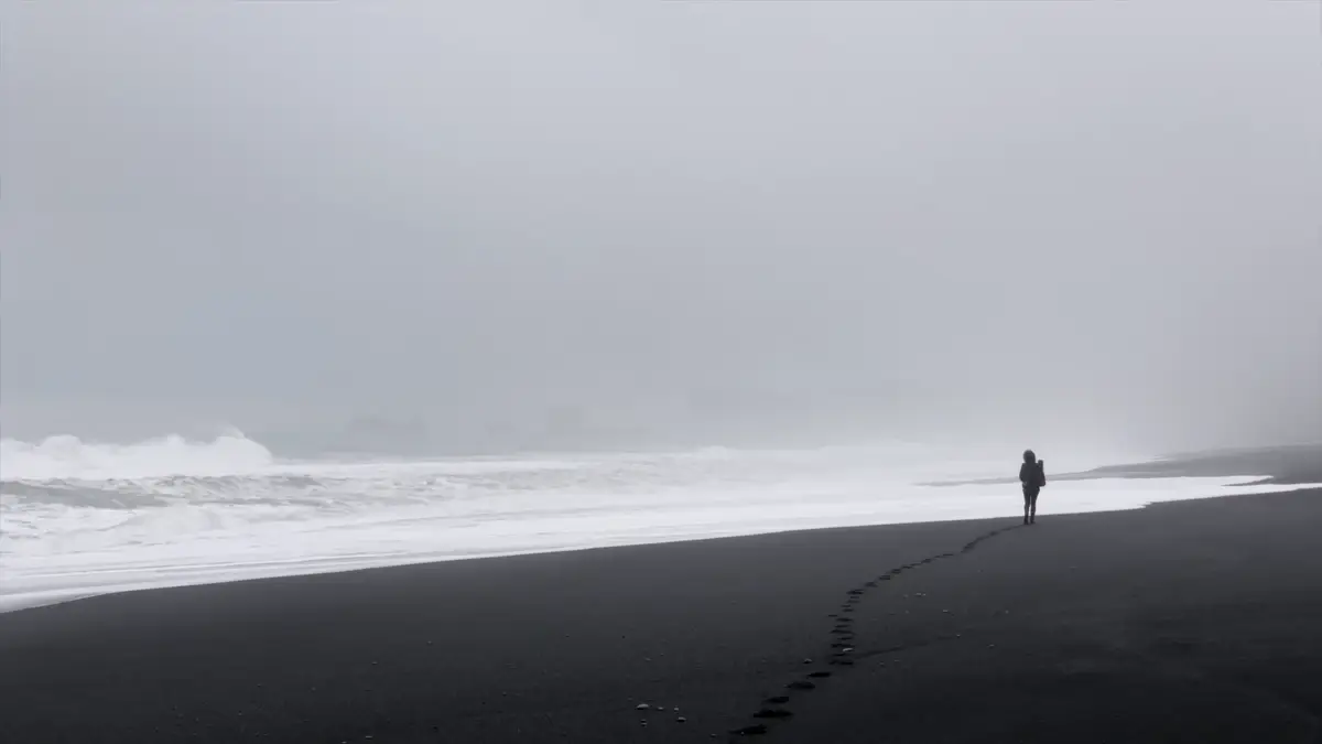 Viajero caminando solo por la playa de arena negra en Islandia, rodeado de niebla y con el mar agitado al fondo. Un destino imprescindible en un road trip en autocaravana.