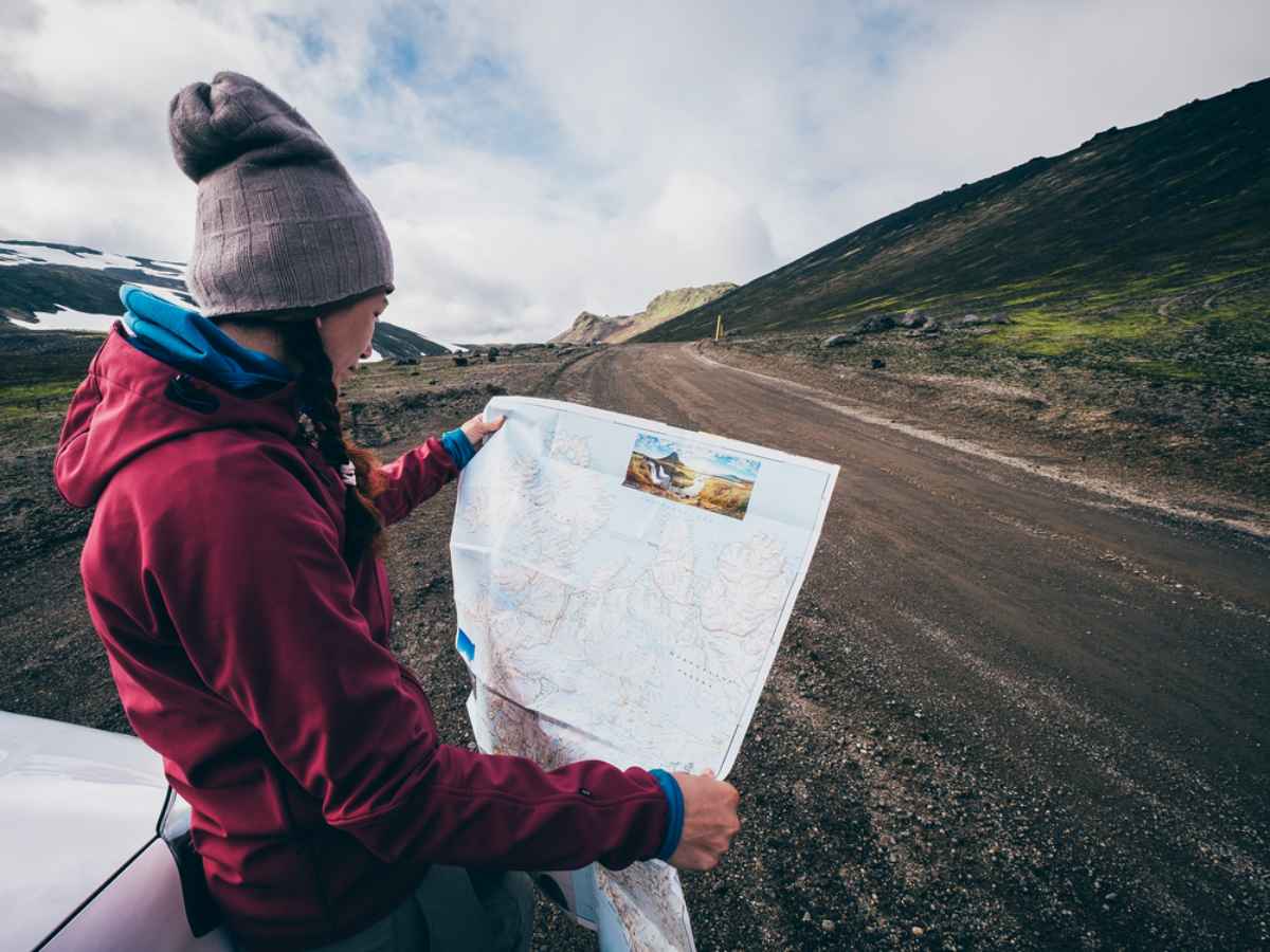 Una mujer sonriente, con gorra y trenzas, consulta una mapa de Islanda mientras su veh&iacute;culo permanece estacionado en la carretera.