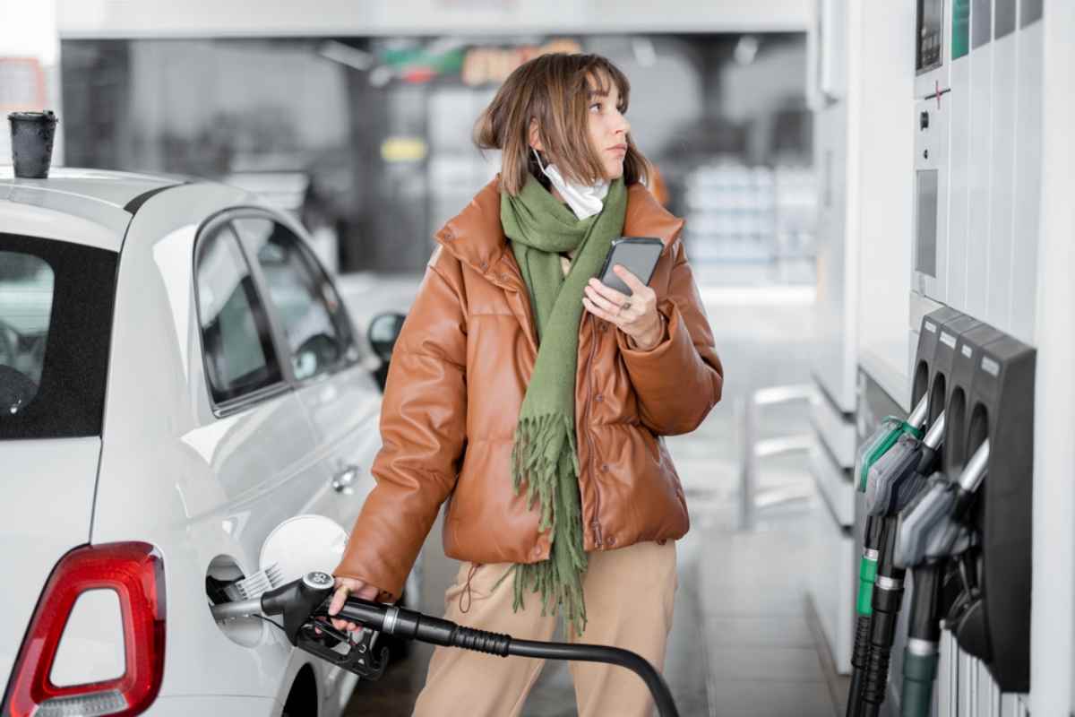 Una chica con abrigo de cuero marr&oacute;n sostiene la manguera en la boca del dep&oacute;sito de su veh&iacute;culo mientras con la otra mano sujeta el m&oacute;vil para calcular el precio de la gasolina en Islandia.
