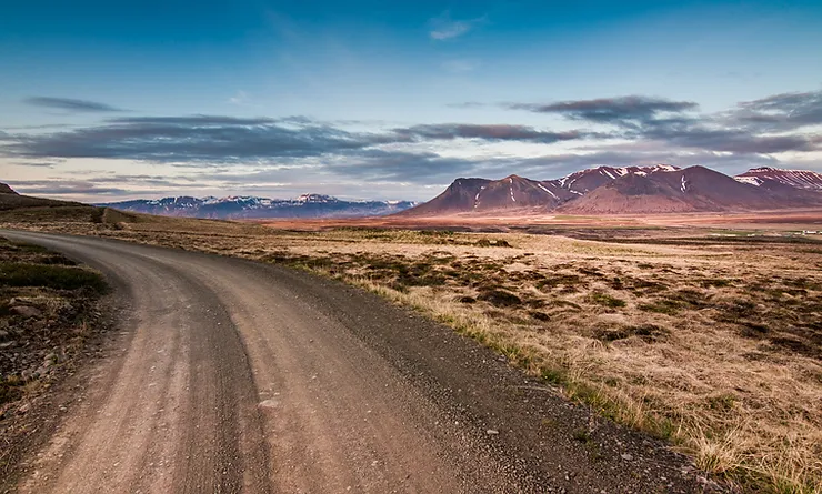 Carretera de grava sinuosa que cruza la tundra abierta y conduce hacia montañas nevadas a lo lejos, bajo un cielo azul con nubes dispersas.