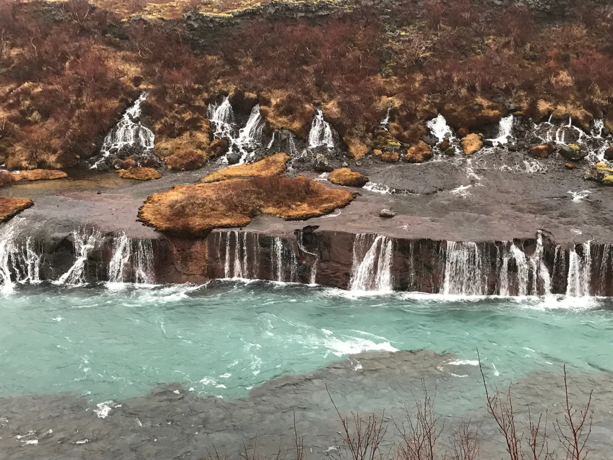 Una cascada islandesa poco empinada durante lo que parecen los últimos resquicios del otoño por el color de la vegetación que la rodea.