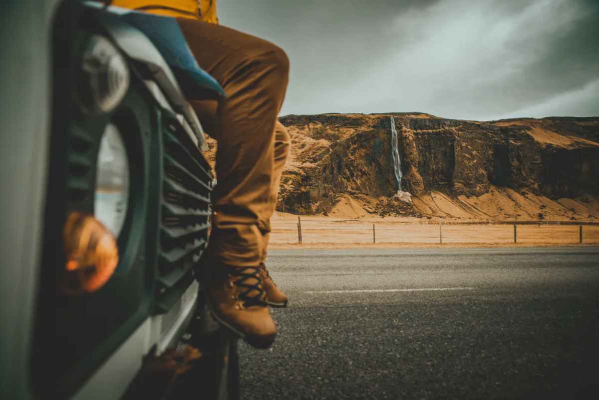 Plano detalle de las piernas de una persona sentada en el capó de su coche estacionado en una carretera islandesa desde donde se aprecia una cascada a