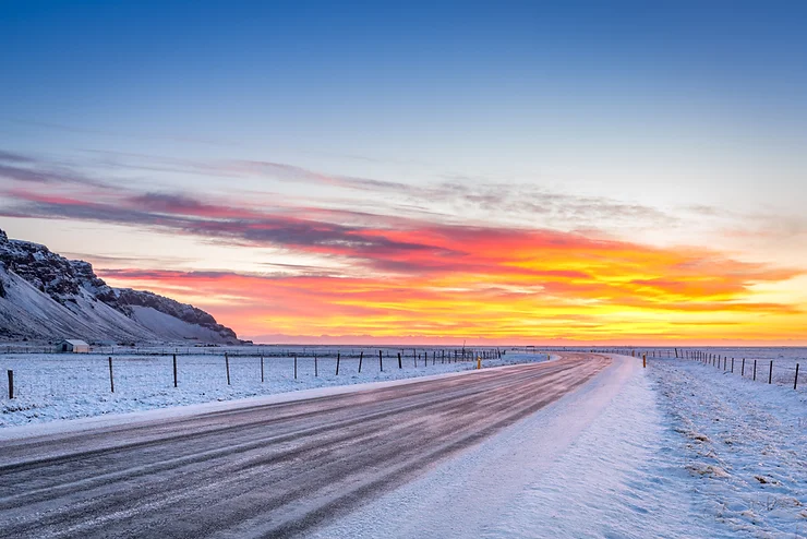 Carretera islandesa cubierta de nieve al atardecer, con el cielo teñido de tonos naranjas y rosas sobre un paisaje invernal